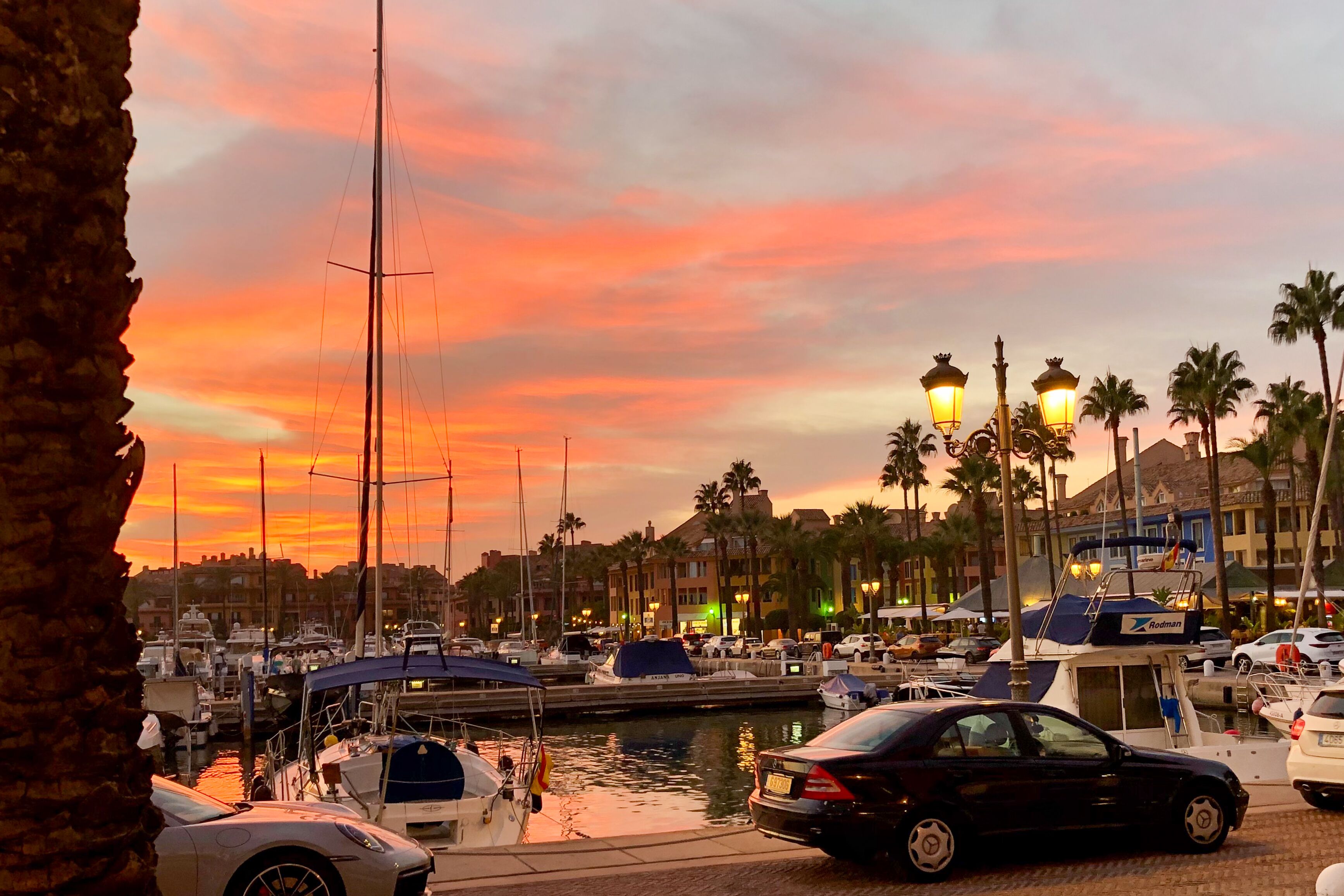 Un port de plaisance avec des bateaux amarrés, une voiture garée et un coucher de soleil en arrière-plan.