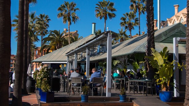 An outdoor café with tables and chairs, surrounded by palm trees and buildings. - Charming apartment / Precioso apartamento en la Marina del Puerto de Sotogrande. (San Roque)