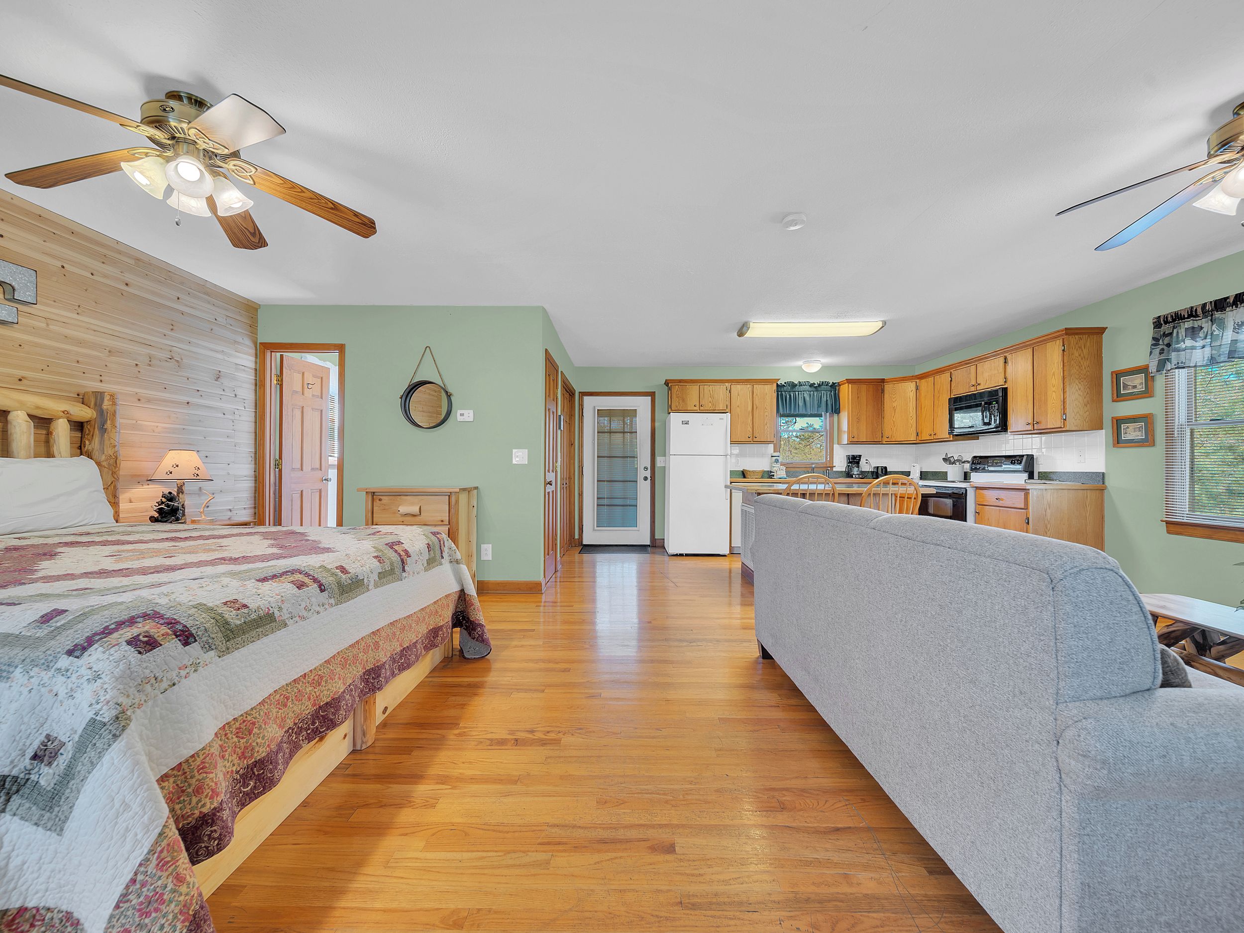 A cozy room with a bed, a sofa, a wooden floor, a ceiling fan, and a kitchen area with wooden cabinets and appliances.