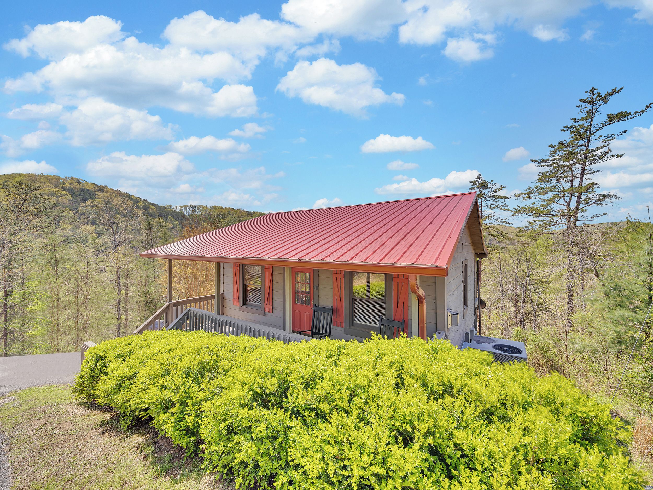 A small cabin with a red roof, surrounded by dense green bushes, set against a backdrop of a forested hillside under a clear blue sky.
