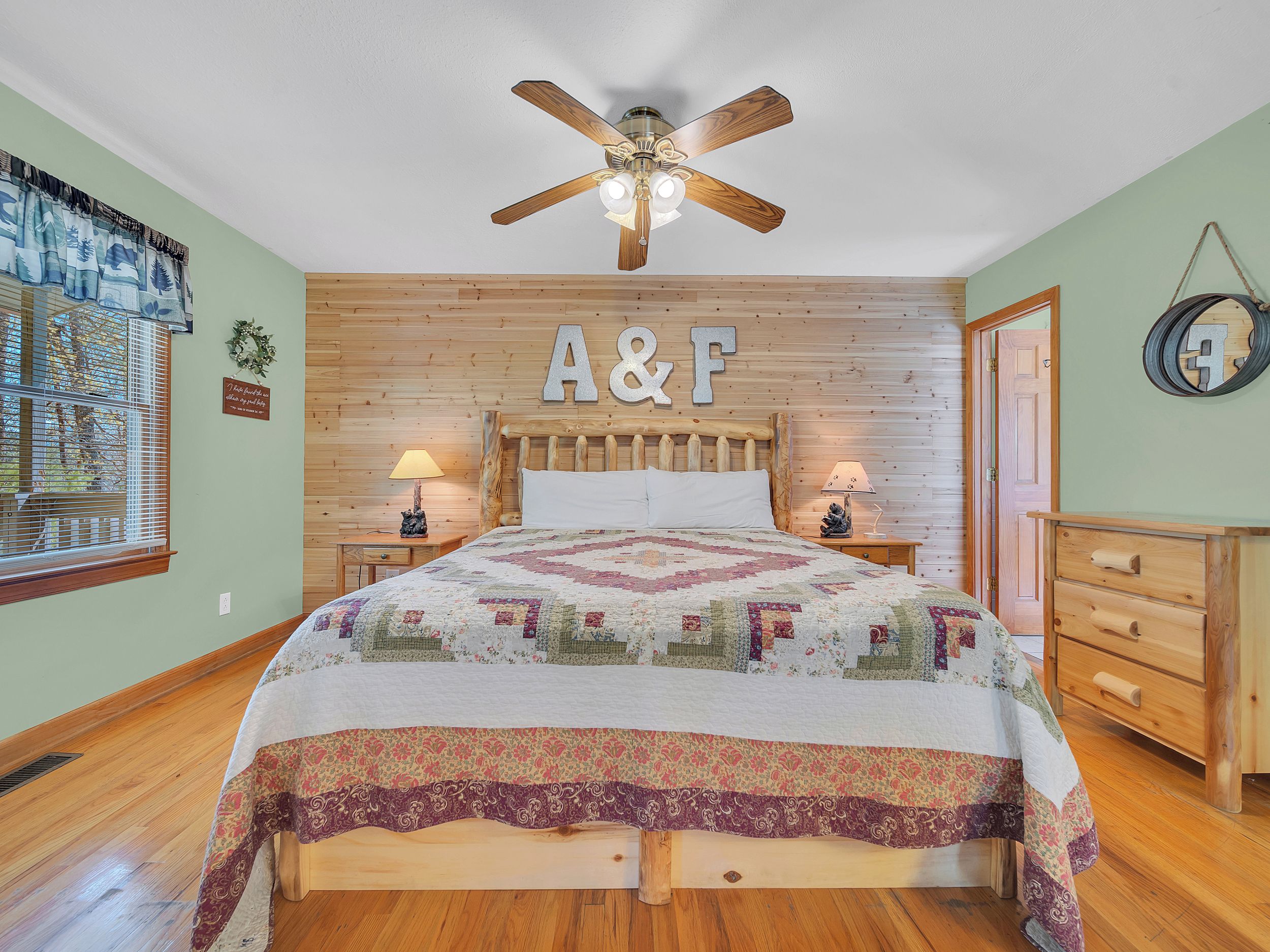 A bedroom with a wooden headboard, a quilted bedspread, two bedside lamps, a ceiling fan, and a wooden dresser.