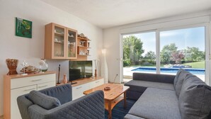 Een moderne woonkamer met een televisie, bank en eethoek. Een groot raam biedt uitzicht op een zwembad en groen.