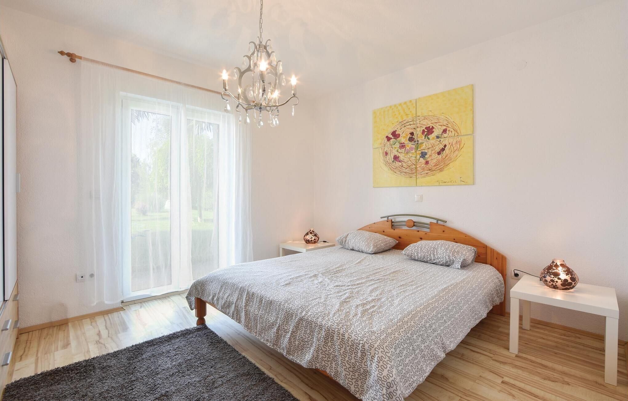 A bedroom with a bed, a chandelier, a painting, and a window with curtains.