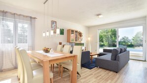 Een moderne woonkamer met een houten eettafel, lichtkleurige stoelen, een grijze bank en een groot raam met uitzicht op groen.