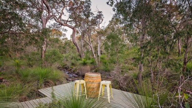 Une terrasse en bois avec deux tabourets et un tonneau, entourée d’une végétation luxuriante et d’arbres.