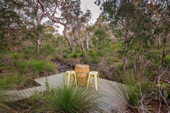 A wooden deck with two stools and a barrel, surrounded by dense vegetation and trees.