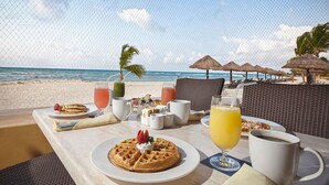 Une table de petit-déjeuner avec des gaufres, du jus de fruits et du café, donnant sur une plage parsemée de parasols et de palmiers.