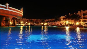 Vue nocturne d’une piscine d’hôtel, avec un grand bassin reflétant les lumières, entourée de bâtiments de l’hôtel et de palmiers.