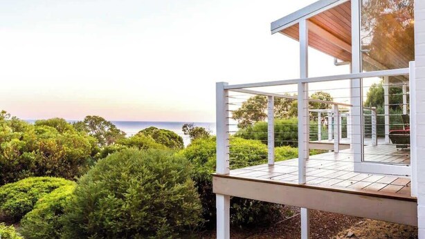 Ett modernt hus med en terrass som vetter ut mot en trädgård och havet.