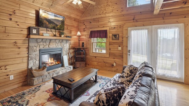 A cozy cabin interior with a stone fireplace, wooden walls, a sofa, and a ceiling fan. - Cozy Cabin|2 King Suites w/Jacuzzis |Near Dollywood (SEVIERVILLE)