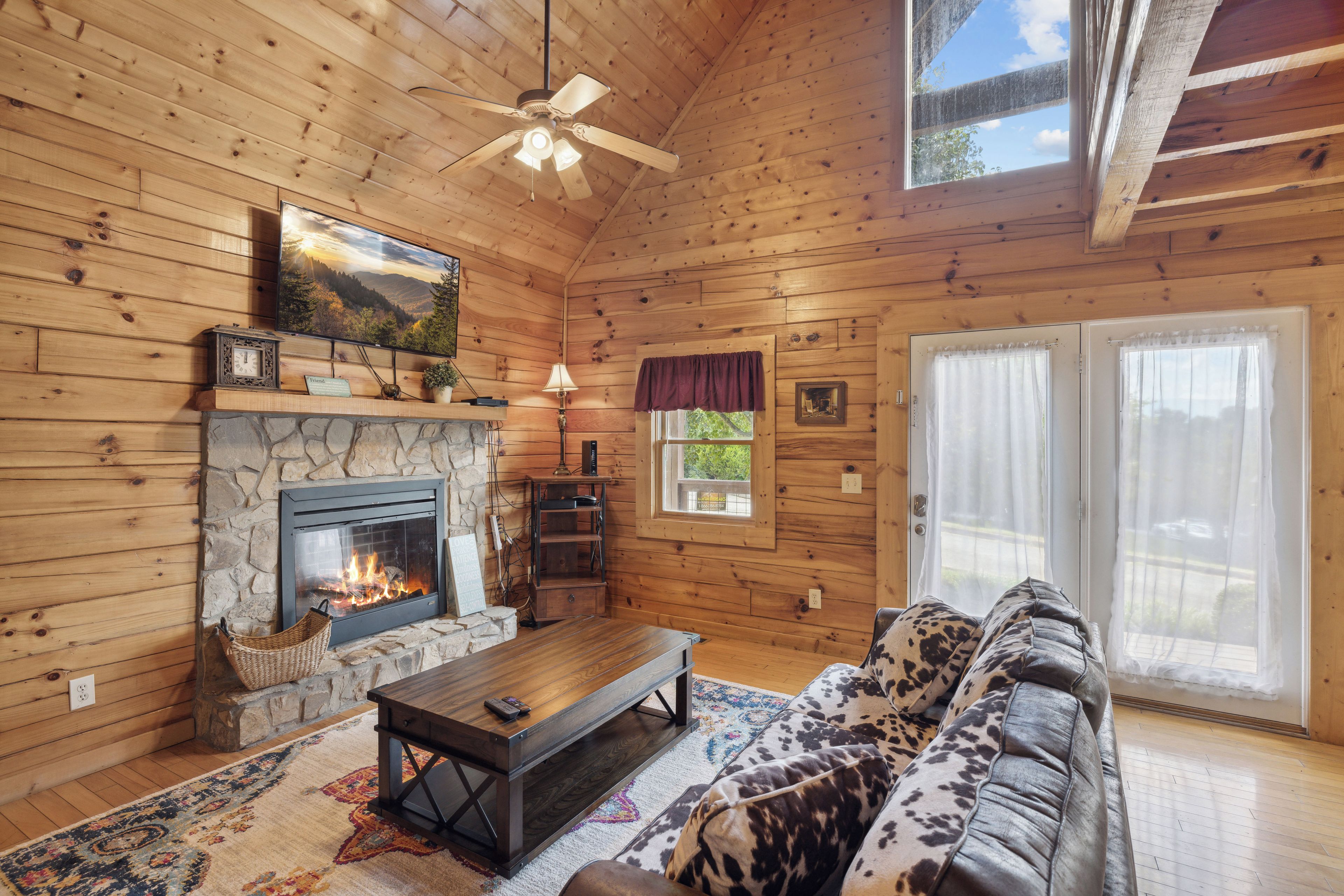 L’intérieur chaleureux d’un chalet avec une cheminée en pierre, un téléviseur fixé au mur, un ventilateur de plafond et une grande fenêtre avec des rideaux.