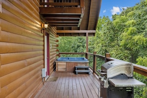 A wooden deck with a hot tub, barbecue grill, and a view of lush greenery. - Cozy Cabin|2 King Suites w/Jacuzzis |Near Dollywood (SEVIERVILLE)