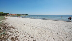 En sandstrand med klart, blåt vand, et par personer i vandet og huse i baggrunden.
