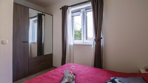 Ein Schlafzimmer mit einem Bett, einem Fenster mit Vorhängen, einem Kleiderschrank und einem Nachttisch mit einer Abstellfläche.