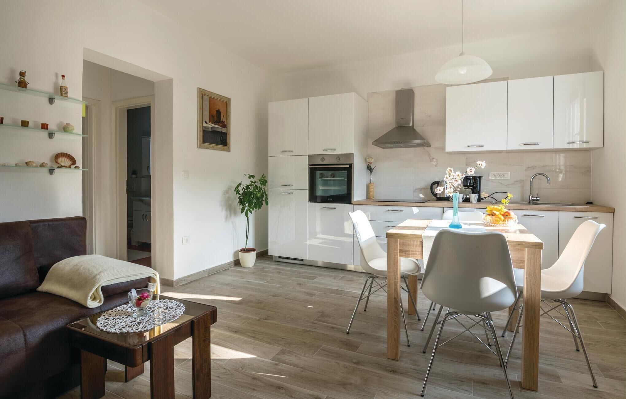 A modern living room with a brown leather sofa, a wooden coffee table, and a dining area with a wooden table and white chairs. A kitchen with white cabinets and stainless steel appliances is visible in the background.