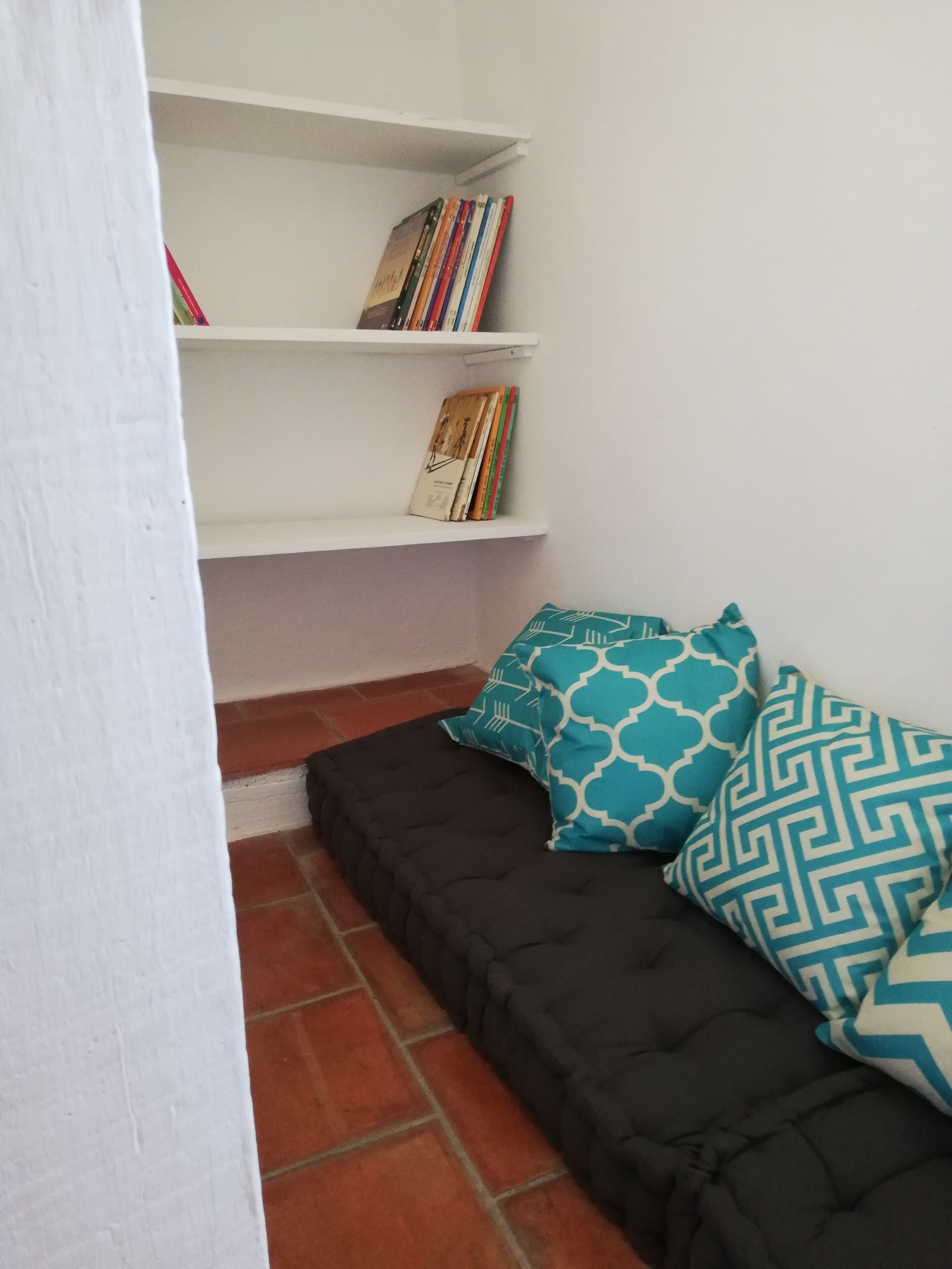 A room with a black sofa, two patterned pillows, a white bookshelf with books, and a tiled floor.