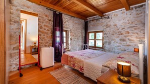 Un dormitorio con paredes de piedra, techo de madera, cama con colcha floral, mesita de noche con lámpara y maleta sobre carrito.