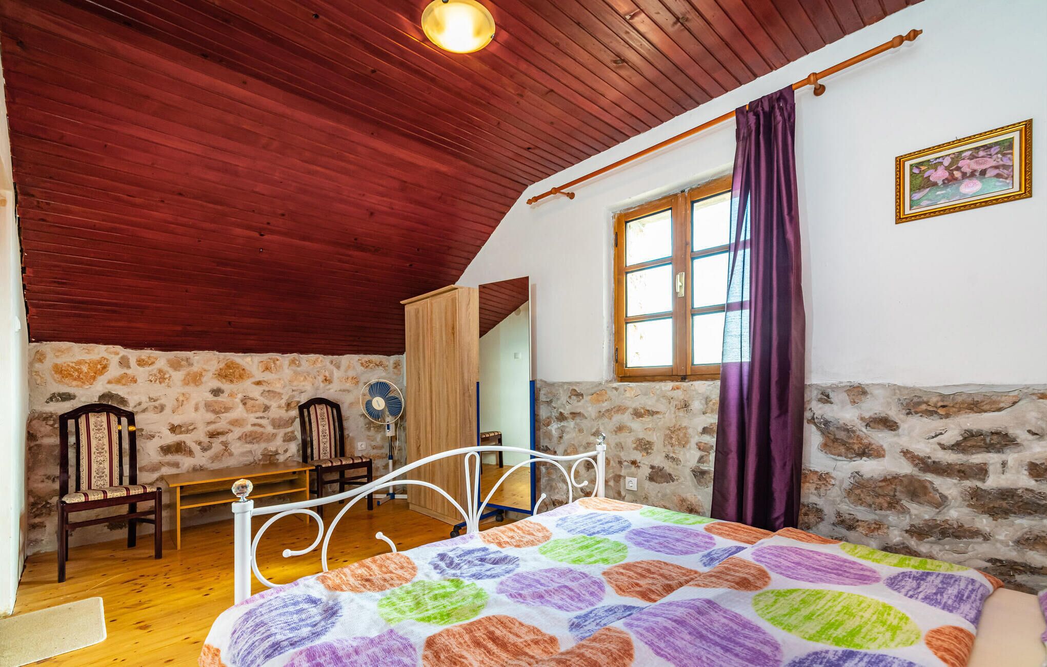Une chambre confortable avec un plafond en bois, des murs en pierre, un lit recouvert d’une courtepointe colorée, une chaise en bois, une petite table et une fenêtre avec des rideaux.