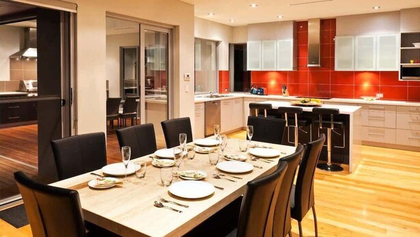 Una cuina moderna amb detalls vermells, una zona de menjador amb taula de fusta preparada per a sis persones, i vista a la sala de dinar veïna.