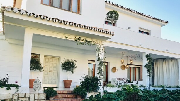 Une maison moderne à deux étages, dotée d’un porche abrité, de plantes en pot et d’un petit jardin.