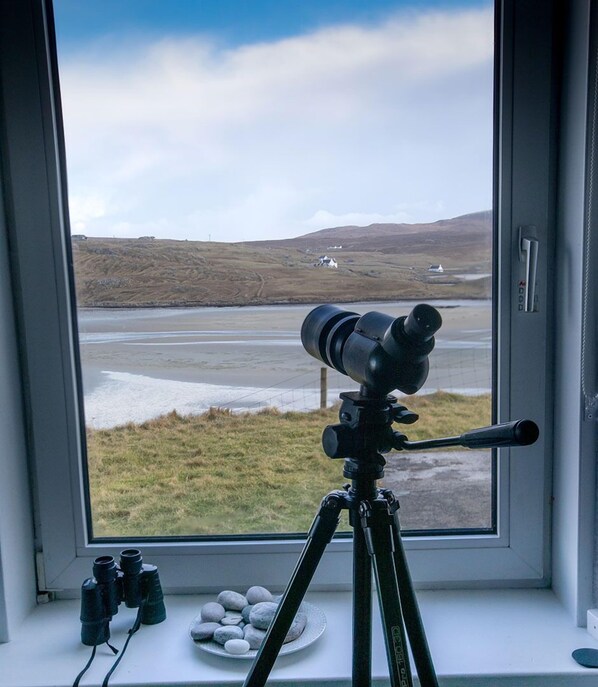 Ein Teleskop auf einem Stativ vor einem Fenster mit Blick auf eine Landschaft, die Hügel und ein Gewässer zeigt.
