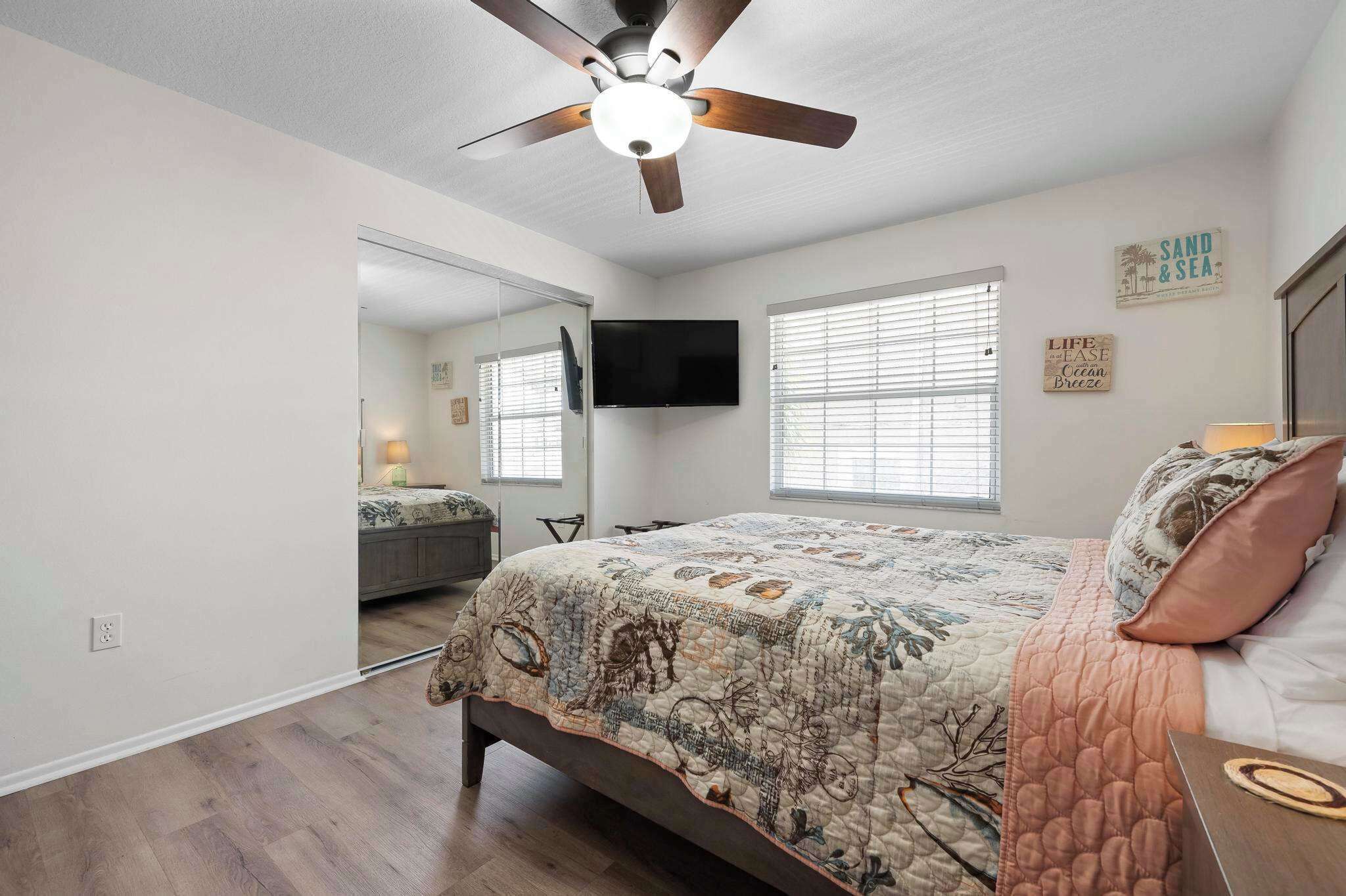 A bedroom with a bed, a ceiling fan, a TV mounted on the wall, and a window with blinds.