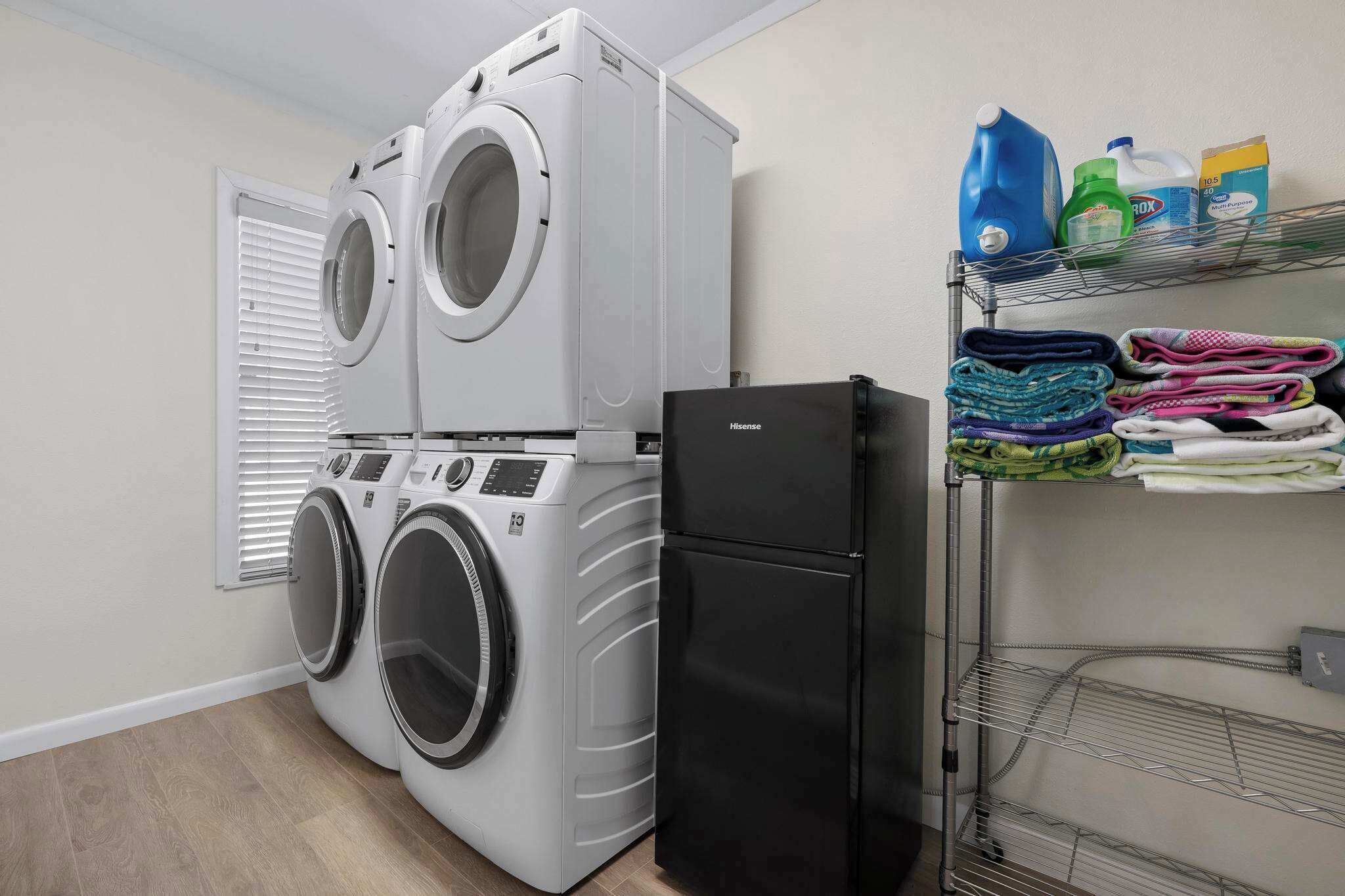 A laundry room with two stacked washing machines, a black refrigerator, and a shelf with folded clothes and cleaning supplies.