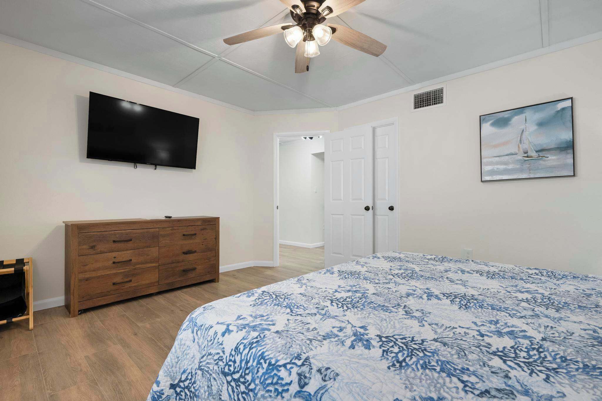 A bedroom with a bed, a TV, a ceiling fan, a dresser, and a door leading to another room.