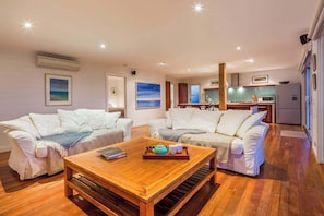 Una sala de estar con dos sofás blancos, una mesa de centro de madera y una cocina al fondo.