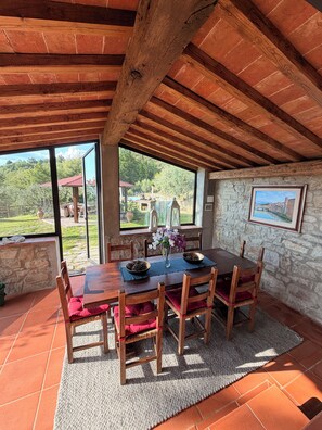 A dining area with a wooden table and chairs, a stone wall, and a view of a garden and buildings outside. - the Casellina (Casa Biondo)
