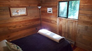 Une petite pièce chaleureuse aux murs en bois, avec un lit recouvert d’une courtepointe bleu foncé, un plaid blanc et deux tableaux encadrés accrochés au mur.