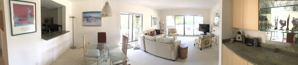 A spacious living room with a glass coffee table, a sofa, and a television. There is a kitchen area with a microwave and a refrigerator.