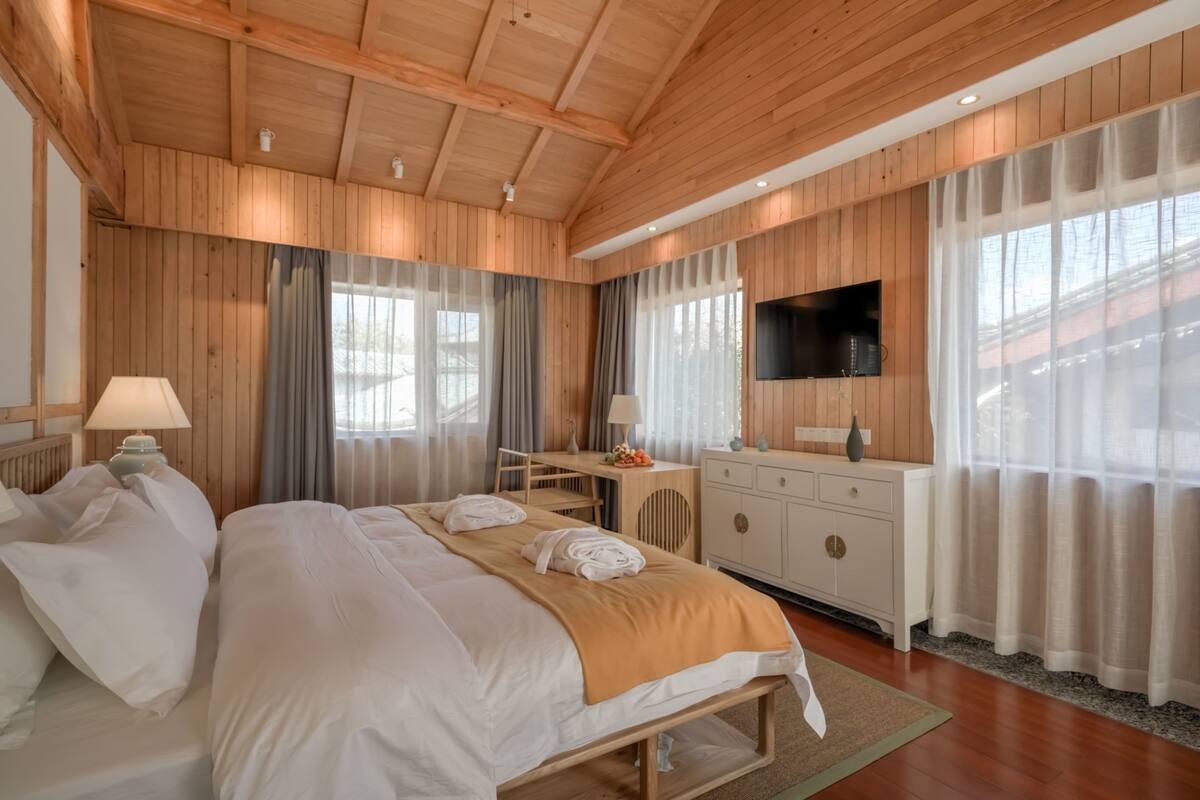 A bedroom with a wooden ceiling, a bed with white linens, a desk with a chair, a TV mounted on the wall, and a window with sheer curtains.