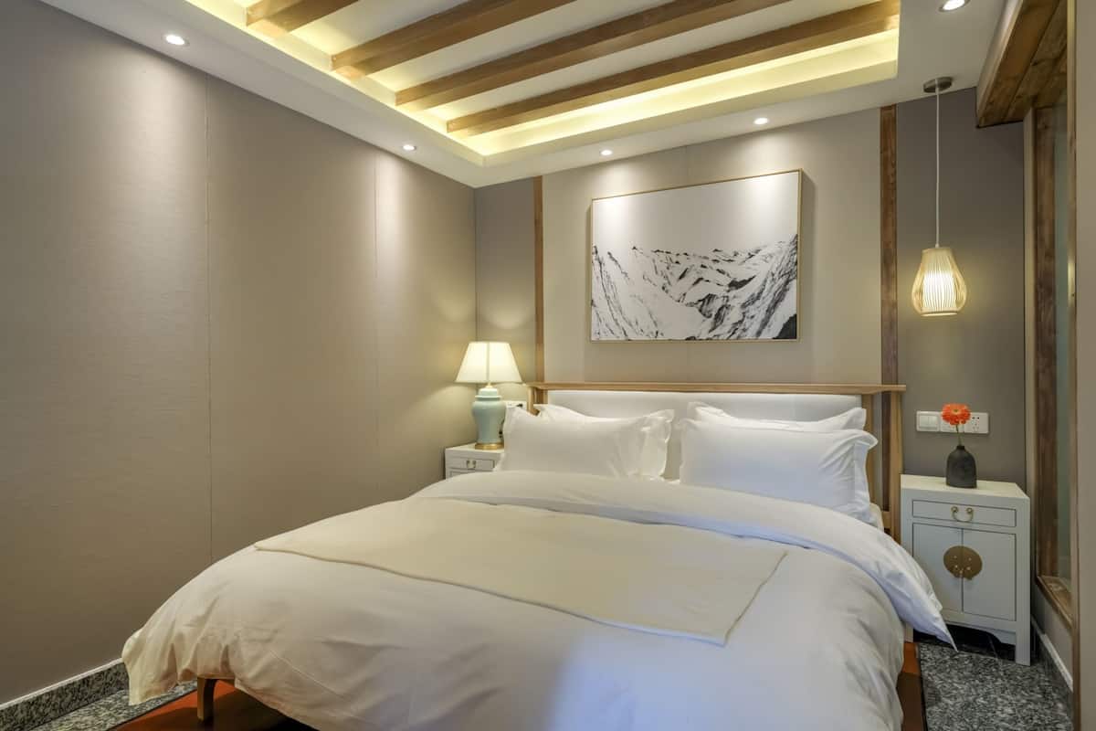 A modern hotel room with a large bed, wooden ceiling beams, a marble artwork, a nightstand with a lamp, and a vase with a flower.