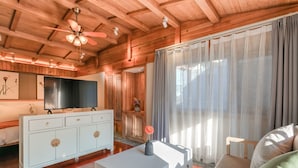 A spacious living room with a wooden ceiling, a large TV on a white cabinet, a sofa, and a window with sheer curtains. - Floral Hotel Lijiang Huazizai (Lijiang)