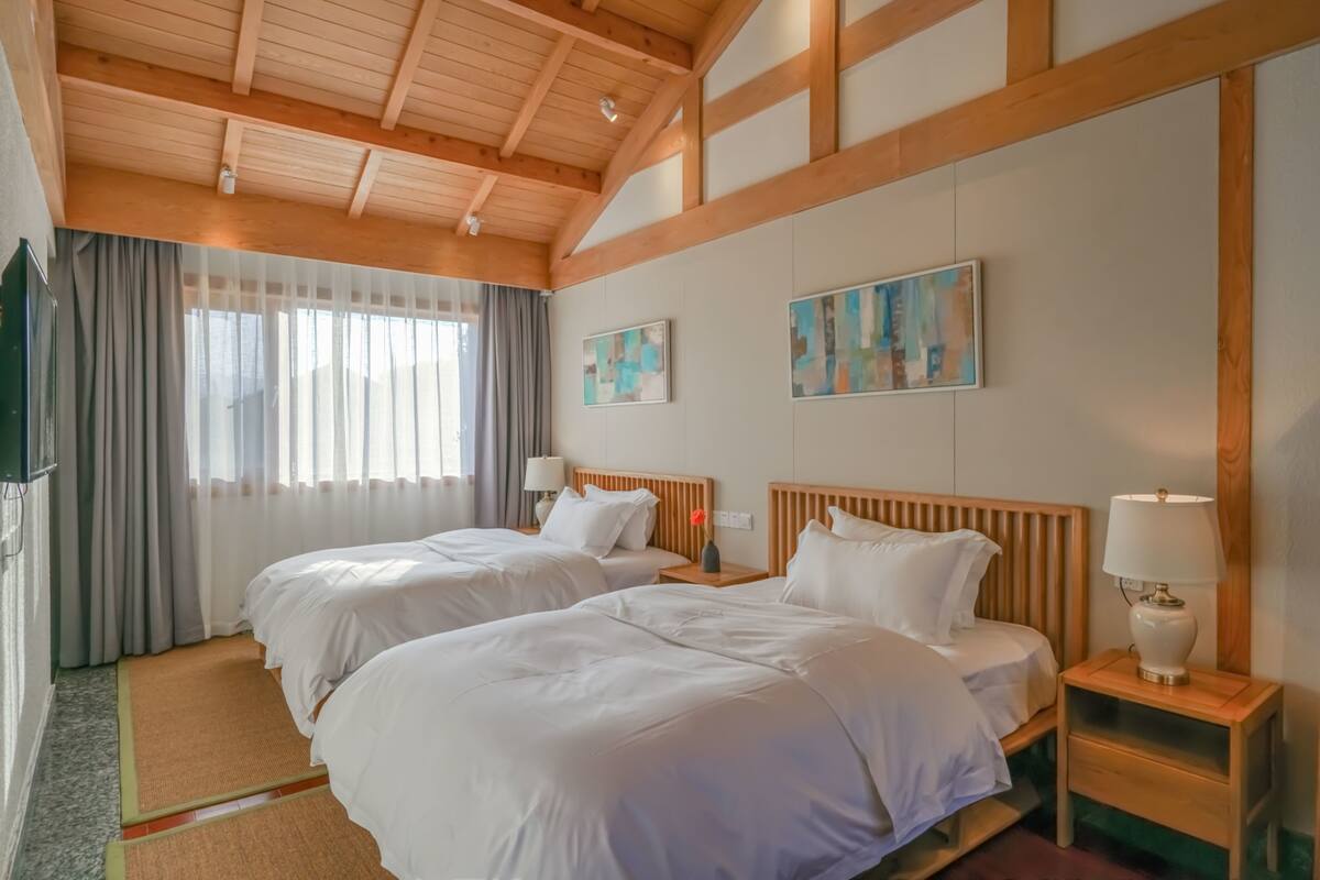 A hotel room with two beds, wooden headboards, a large window with curtains, and artwork on the walls.
