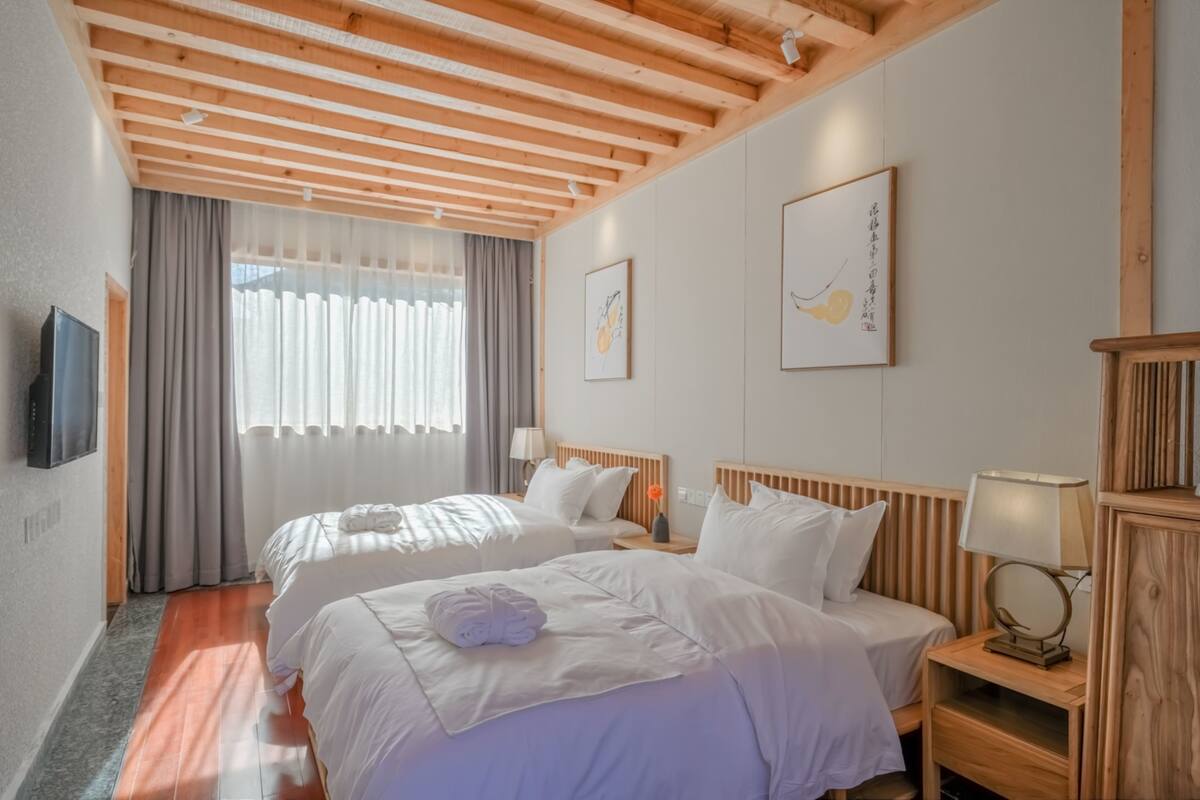A hotel room with two beds, wooden headboards, a flat-screen TV, a window with curtains, and artwork on the walls.