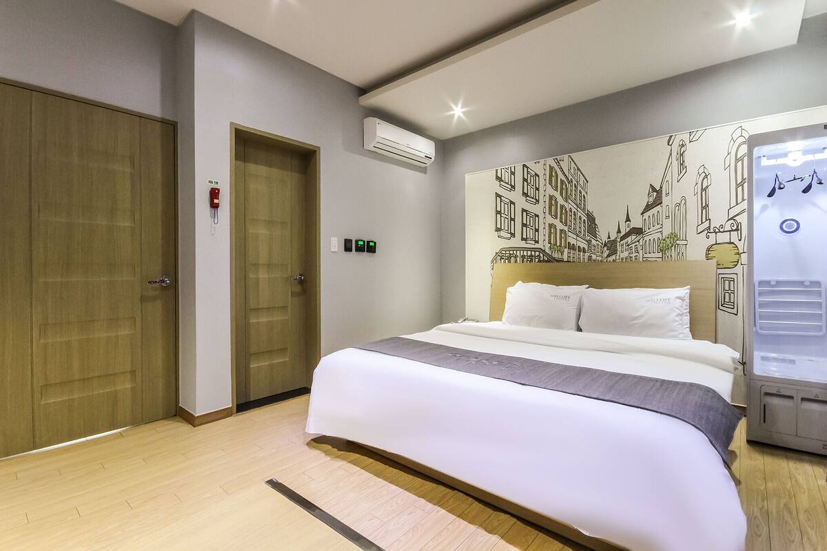 Habitación de hotel con una cama grande, un armario de madera, un aire acondicionado instalado en la pared y un mural de un paisaje urbano.