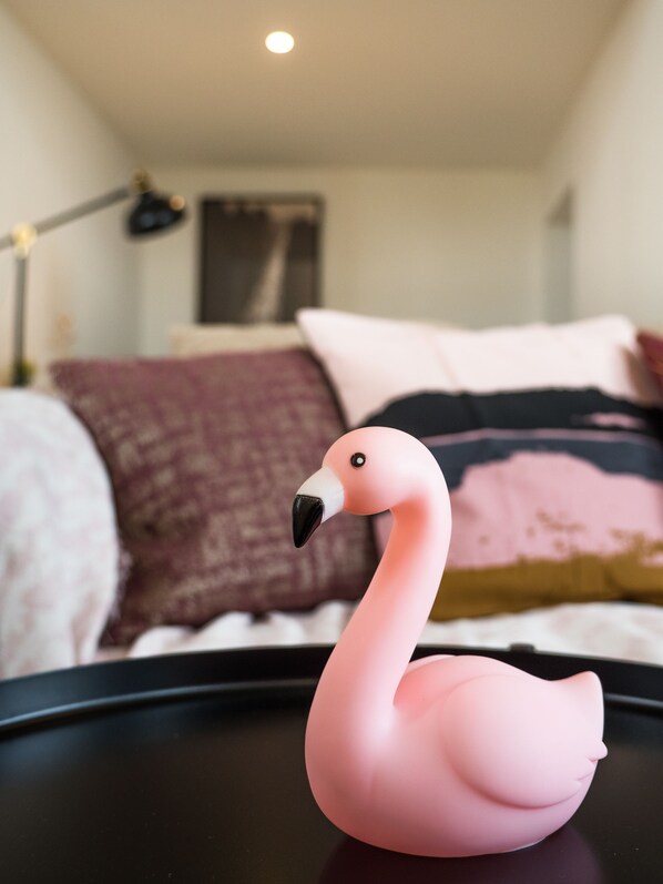 Eine rosa Flamingo-Figur auf einer schwarzen Oberfläche in einem Zimmer mit Bett, Kissen und einer Lampe.
