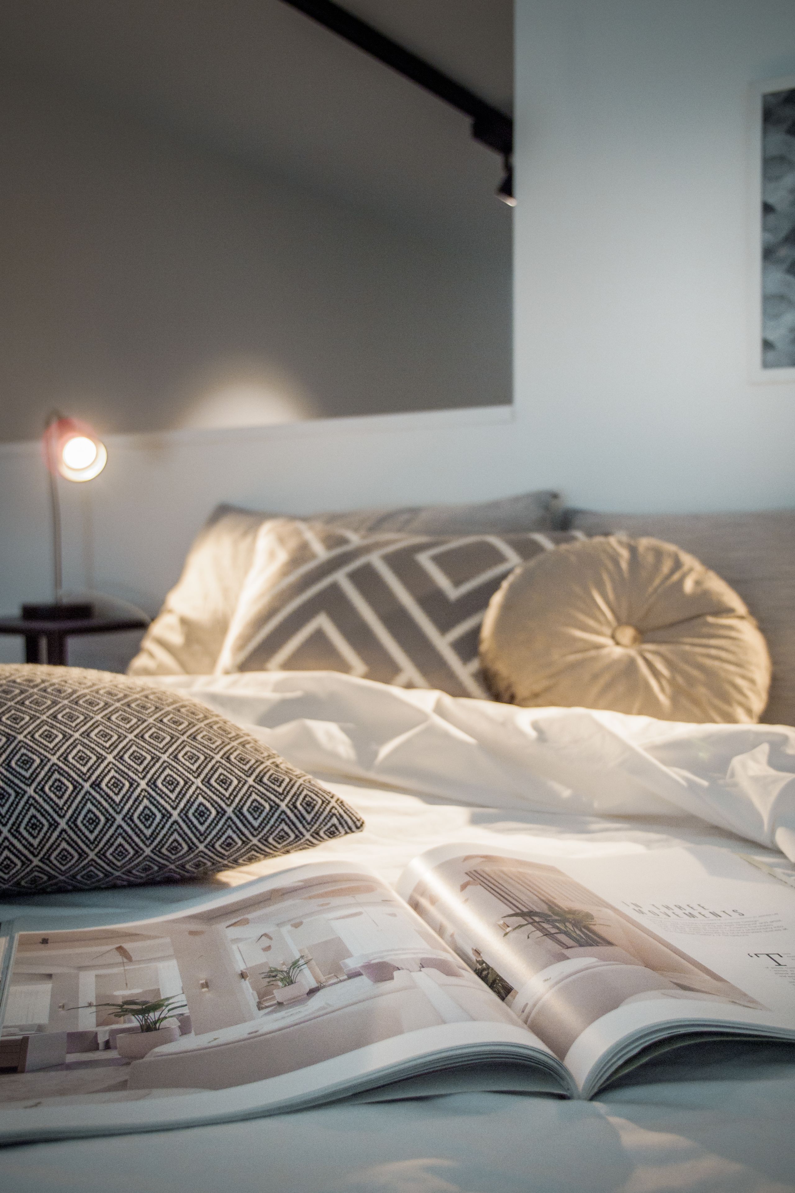 Un lit bien fait, agrémenté d’oreillers à motifs, une lampe de chevet et un magazine ouvert posé sur le lit.