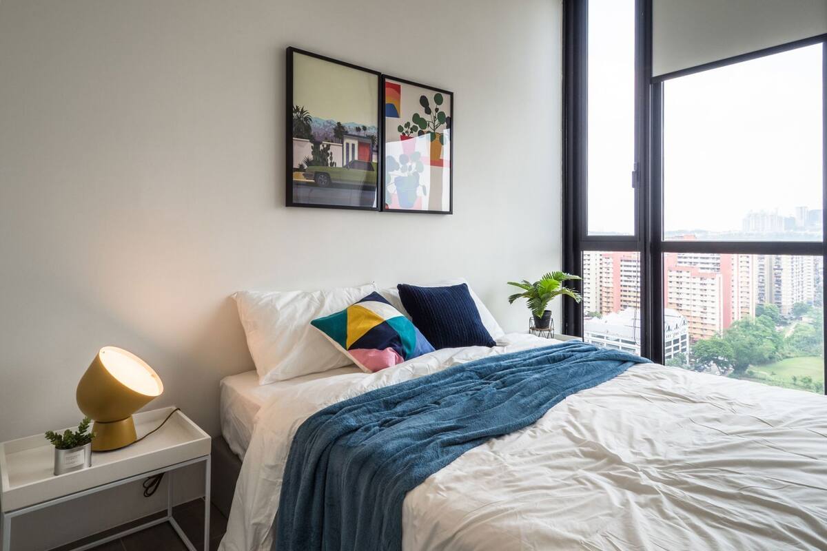 A bedroom with a bed, pillows, a bedside lamp, a small plant, and a window with a city view.