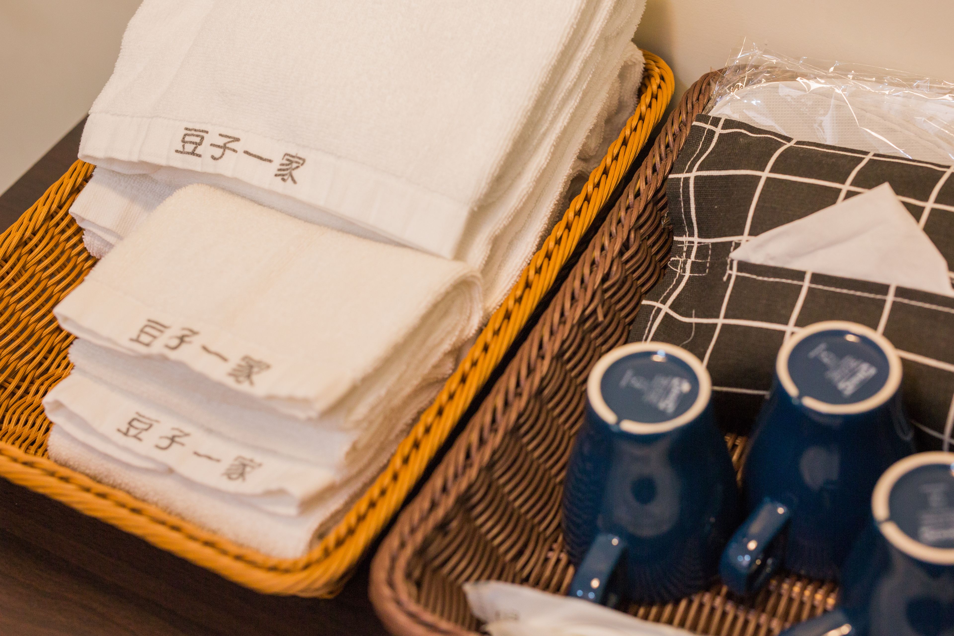 A wicker basket containing folded white towels with Japanese text, a black and white checkered pillow, and blue mugs.