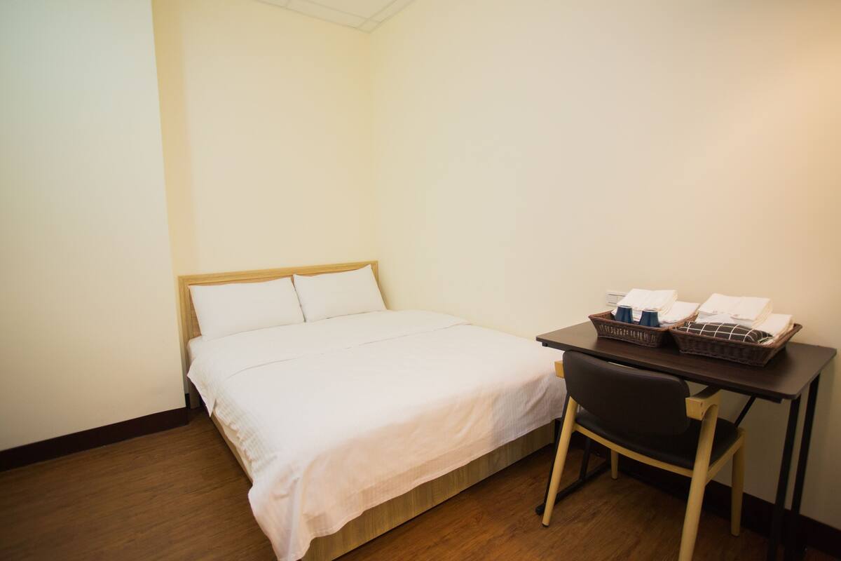 Ein Hotelzimmer mit einem Bett, einem Schreibtisch mit Stuhl und einem kleinen Tisch mit einer Korbablage für Handtücher und Tassen.