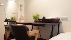 A compact hotel room with a bed, a desk with a chair, a small potted plant, a hairdryer, and a basket on the desk.