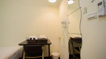 A small, compact room with a single bed, a desk with a chair, a telephone, and a waste bin.