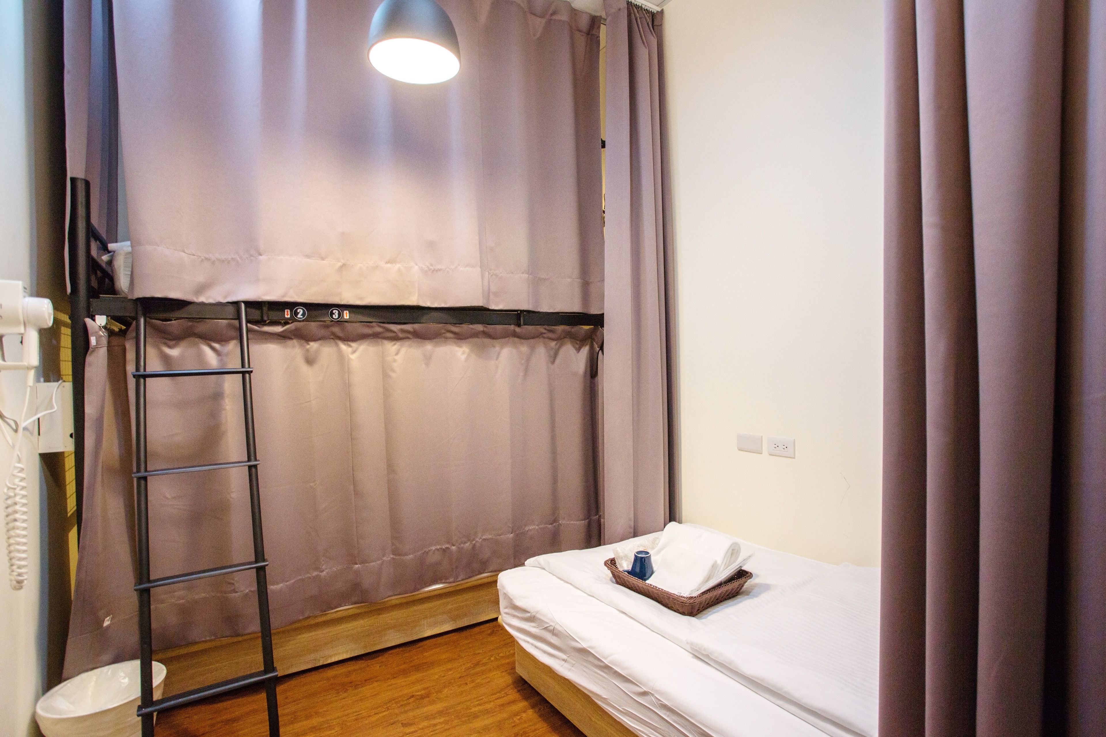 Une petite chambre avec un lit simple, un plancher en bois, un lit blanc, une petite table avec une bouteille, une échelle et un rideau.