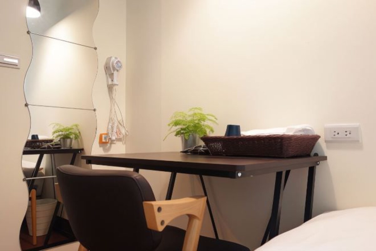 A compact hotel room with a small desk, a chair, a bed, and two potted plants.