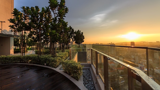 Terraza en la azotea con barandilla de vidrio, árboles y vistas a un paisaje urbano al atardecer.