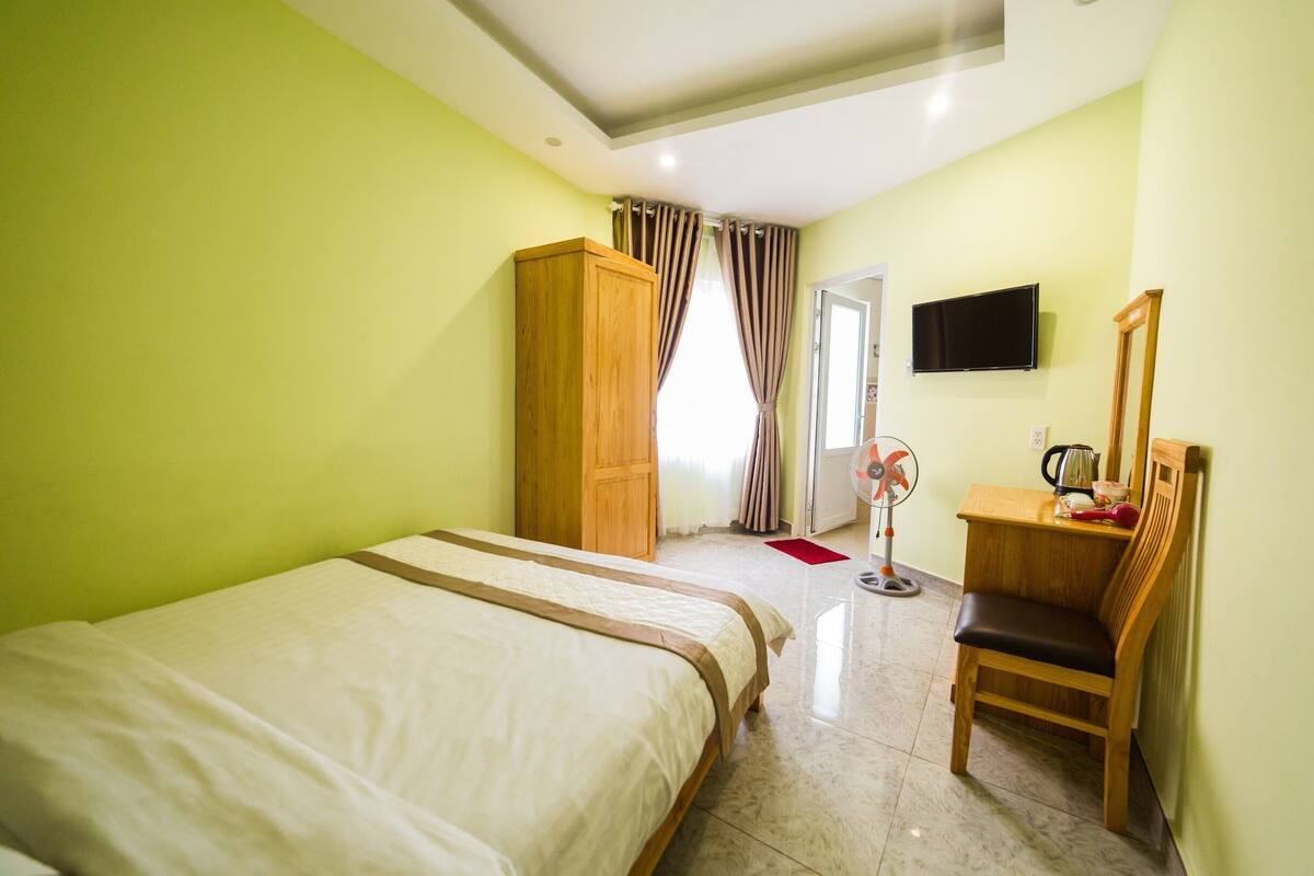 A hotel room with a bed, a wooden wardrobe, a desk with a chair, a television, and a fan.