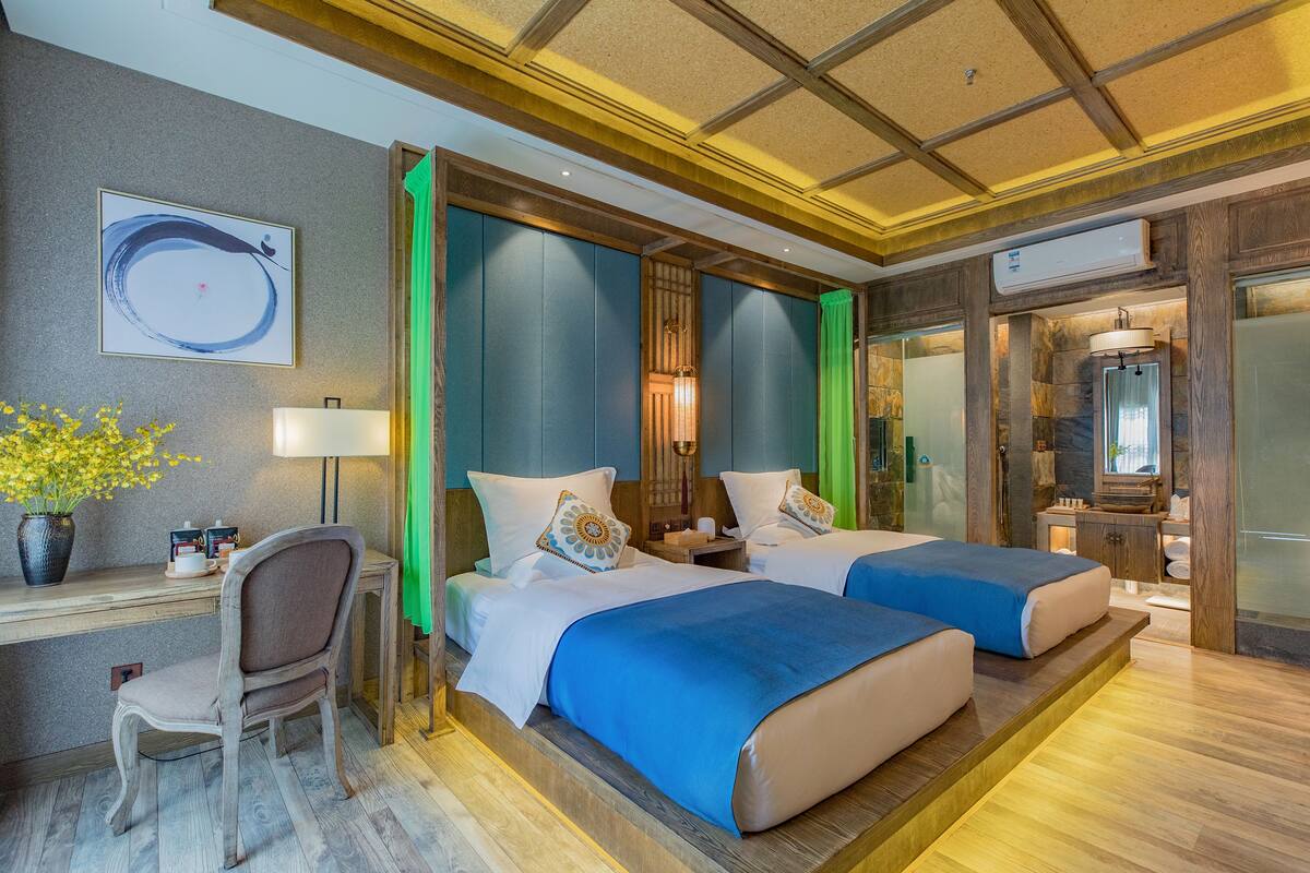 Hotelkamer met twee bedden, een bureau, een stoel, een kamerplant en een badkamer zichtbaar door een glazen deur.